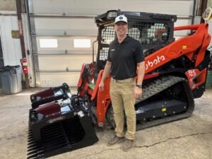 905x0_s3-71357-M-LANO-CAD-26-1jpg - Machines Wiki Man in a black polo and khaki pants standing beside a red skid-steer loader with a large black grapple attachment in a workshop.