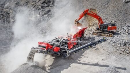 Red rock-crushing and conveying machine at a dusty quarry slope in operation.