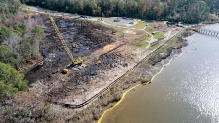 Aerial view of a waterfront construction site with a tall yellow crane, muddy ground, and a yellow containment boom along the lake shore.
