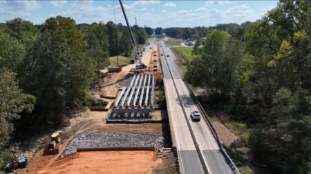 Bird's-eye view of a road construction site: a crane and steel beams beside a highway, with trees on both sides.