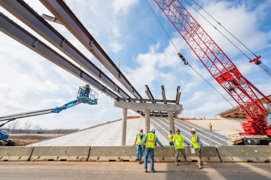 905x0_s3-71377-S-041_26-CR-1jpg - Machines Wiki Construction crew installs large steel beams overhead for a bridge or overpass, with a red crane in the background.