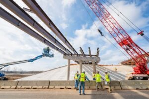 Construction crew installs large steel beams overhead for a bridge or overpass, with a red crane in the background.