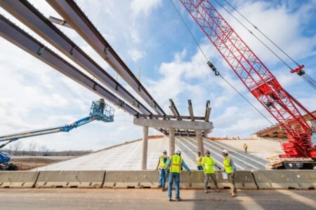 Construction crew installs large steel beams overhead for a bridge or overpass, with a red crane in the background.