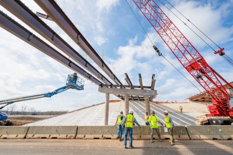 Construction crew installs large steel beams overhead for a bridge or overpass, with a red crane in the background.
