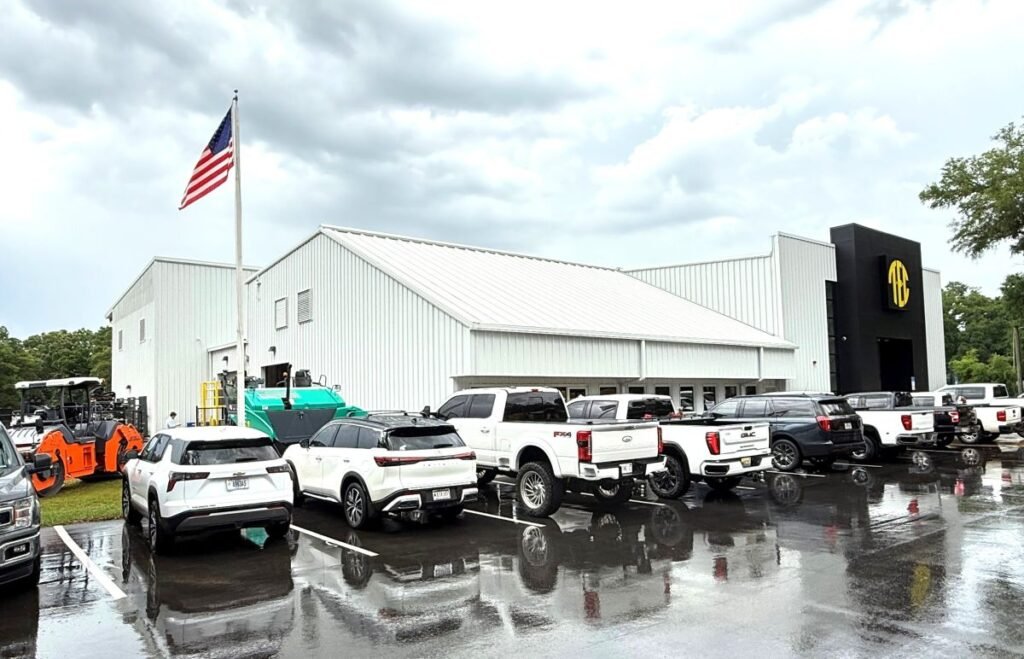 905x0_s3-71379-S-TEC-PEN-5-26-1jpg - Machines Wiki American flag atop a white metal building with a row of white pickup trucks parked on a wet lot, after rain.