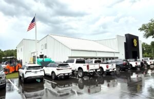 American flag atop a white metal building with a row of white pickup trucks parked on a wet lot, after rain.