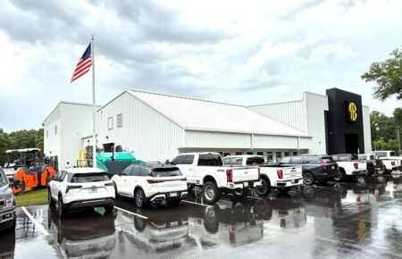 American flag atop a white metal building with a row of white pickup trucks parked on a wet lot, after rain.