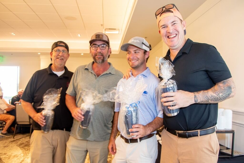 905x0_s3-71385-S-DOBBS-GOLF-26-1jpg - Machines Wiki Four men pose together indoors, each holding a wrapped tumbler gift and smiling at the camera during a casual event.