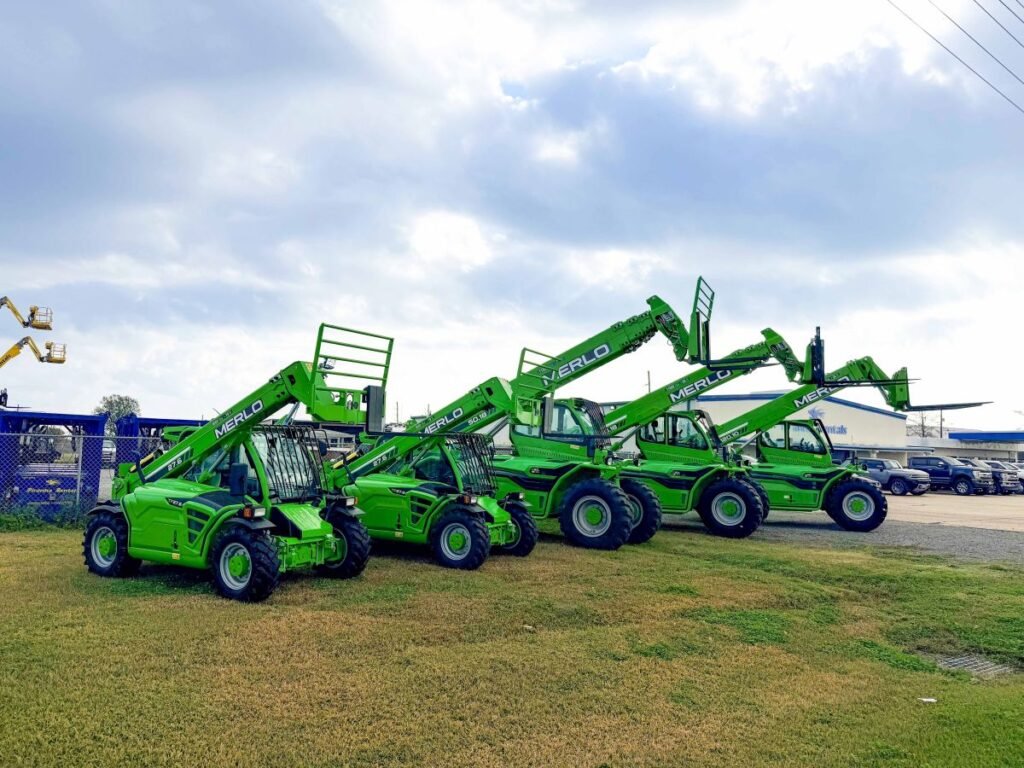 Lineup of bright green Merlo telescopic handlers with extended booms parked on a grassy lot under a cloudy sky