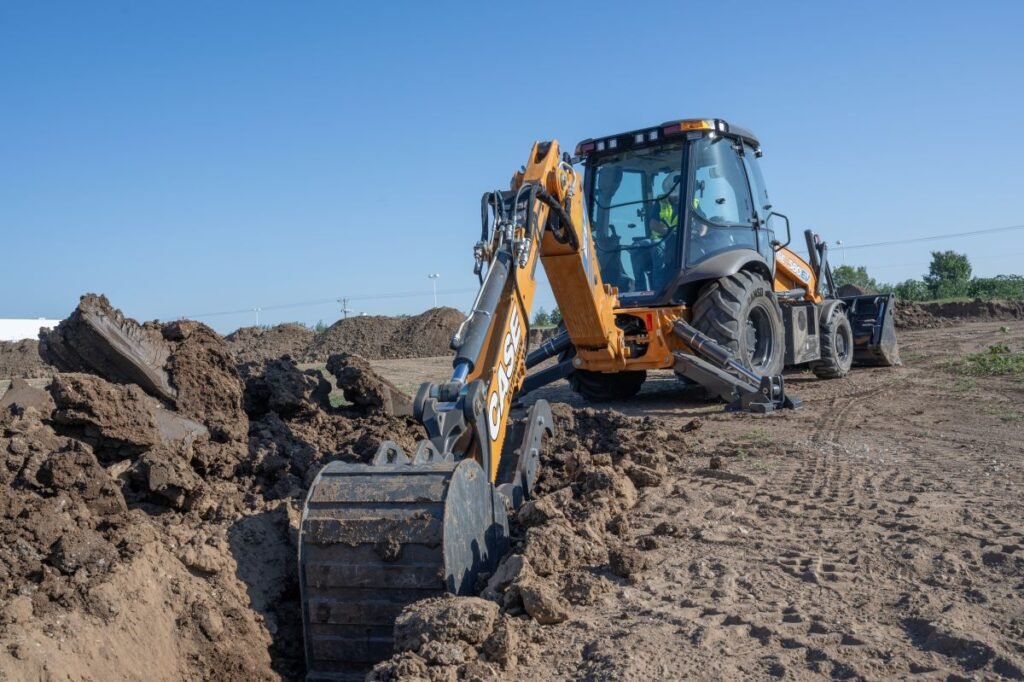 905x0_s3-71405-MNSW-EE-CASE-CE-580EV-1jpg - Machines Wiki Backhoe loader digger moving soil at a construction site with dirt mounds and clear blue sky behind it.