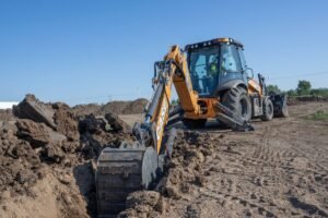 Backhoe loader digger moving soil at a construction site with dirt mounds and clear blue sky behind it.