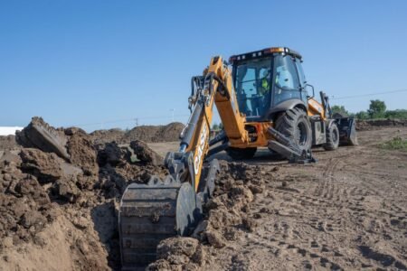 Backhoe loader digger moving soil at a construction site with dirt mounds and clear blue sky behind it.