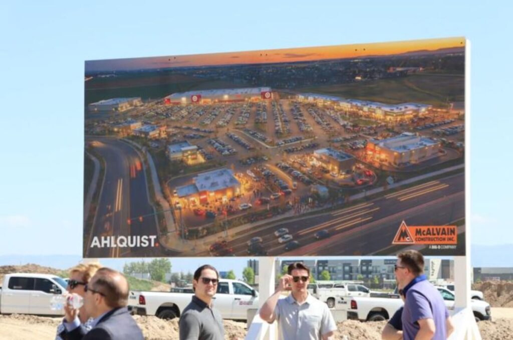 Aerial billboard showing a planned shopping center, with AHLQUIST and McALVAIN Construction logos, at a construction site with people and trucks nearby.