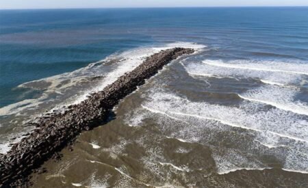 A rocky breakwater extends from the shore into the sea, with waves crashing along its sides beneath a blue ocean sky.