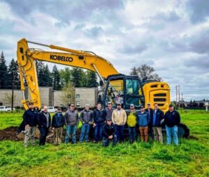 Group of construction workers posing in front of a large yellow Kobelco excavator on a grassy lot with a building in the background, outdoors.