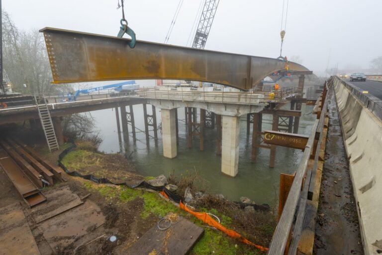 Large rusted steel beam being hoisted by crane over a river while a bridge is built nearby. Construction equipment and a roadway are visible in foggy conditions.