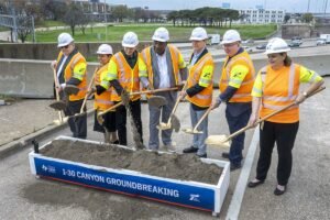 Group of officials and workers in hard hats and orange vests shovel dirt at an I-30 Canyon Groundbreaking ceremony, standing in a parking lot.