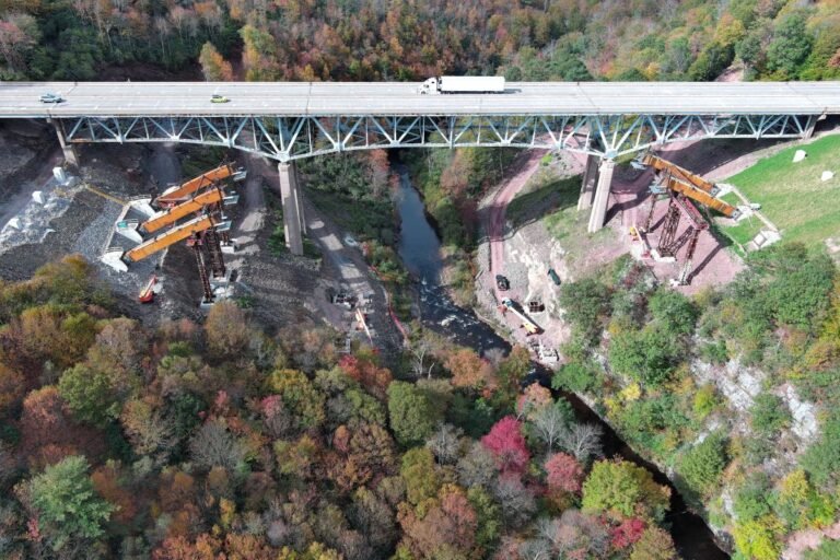 Aerial view of a large bridge under construction over a forested gorge, with cranes and construction equipment on the riverbank side.