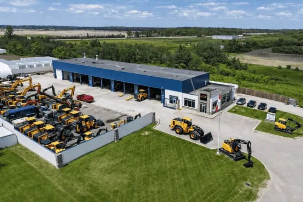 905x0_s3-71418-RTI-Office_-768x512_webpjpg - Machines Wiki Aerial view of a heavy equipment dealership with rows of yellow construction machines in a fenced lot beside a blue-roofed building.