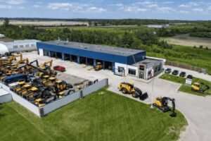 Aerial view of a heavy equipment dealership with rows of yellow construction machines in a fenced lot beside a blue-roofed building.