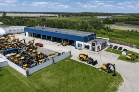 Aerial view of a heavy equipment dealership with rows of yellow construction machines in a fenced lot beside a blue-roofed building.
