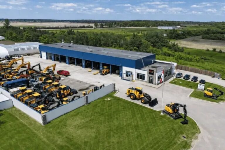 Aerial view of a heavy equipment dealership with rows of yellow construction machines in a fenced lot beside a blue-roofed building.