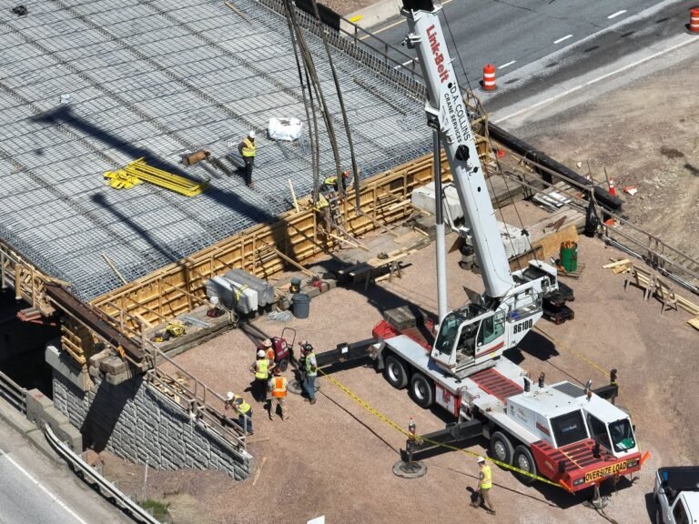 Aerial view of a construction site with a tall crane lifting materials over a reinforced concrete area, workers in vests and helmets nearby.