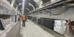 Inside a curved underground tunnel under construction; workers in high-visibility vests and hard hats stand near stacked barriers, pipes, and equipment.