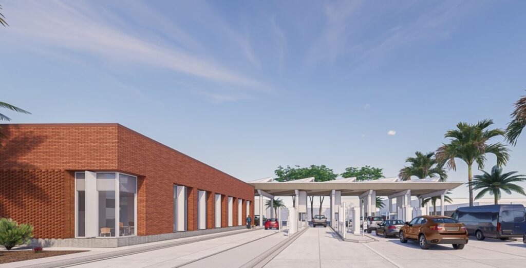 Modern brick building with tall glass windows on the left, a covered drop-off area, and parked cars under a blue sky.
