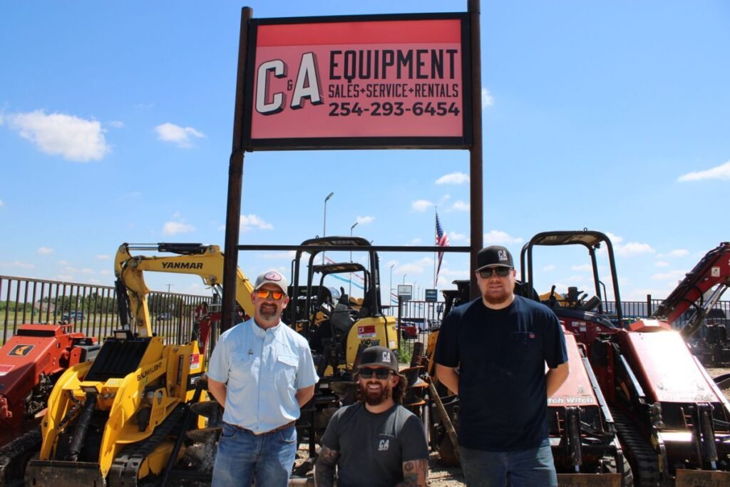 Three men pose in front of a CA Equipment rental lot with yellow and red construction machines behind them.