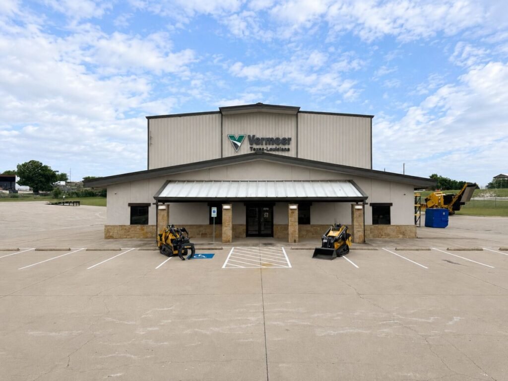 Front of a Vermeer facility with a beige facade, metal awning, and two yellow skid-steer loaders parked near the entrance in a large empty lot.