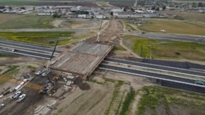 Aerial view of a bridge under construction spanning a highway, with cranes, scaffolding, and construction equipment on site.