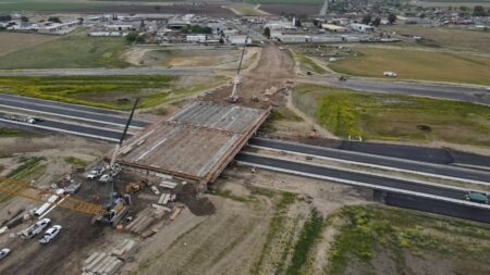 Aerial view of a bridge under construction spanning a highway, with cranes, scaffolding, and construction equipment on site.