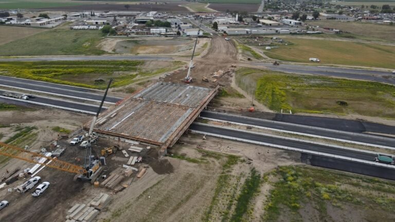 Aerial view of a bridge under construction spanning a highway, with cranes, scaffolding, and construction equipment on site.