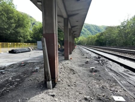 Railroad platform under construction with tracks to the right, yellow safety fence, and a leaning damaged support column amid gravel and construction materials nearby.