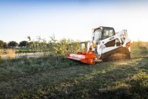 Bobcat-T650-Compact-Track-Loader-3_redjpg - Machines Wiki White skid-steer loader with an orange brush mower attached, clearing tall grass in a rural field at sunset/late afternoon sun.