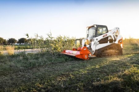 Bobcat-T650-Compact-Track-Loader-3_redjpg - Machines Wiki White skid-steer loader with an orange brush mower attached, clearing tall grass in a rural field at sunset/late afternoon sun.
