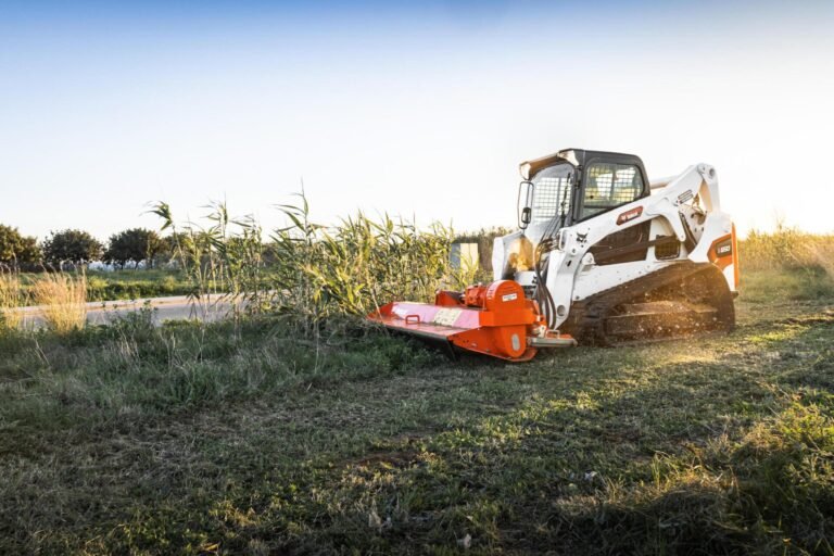 White skid-steer loader with an orange brush mower attached, clearing tall grass in a rural field at sunset/late afternoon sun.