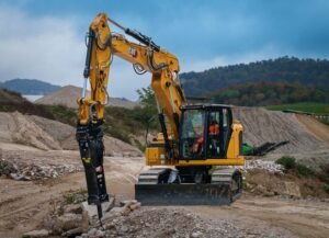 Yellow CAT excavator with hydraulic breaker at a rocky construction site, operator visible in cab