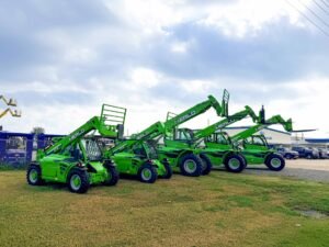 Dealership-pic-3jpg - Machines Wiki Row of green Merlo telehandlers parked on a grassy lot under a cloudy sky with a chain-link fence in the background.
