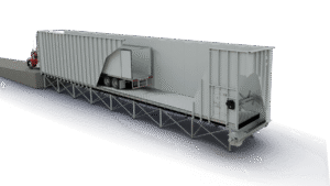 Red semi truck at a loading dock beside a long gray warehouse, with a trailer entering a loading bay under a metal platform.