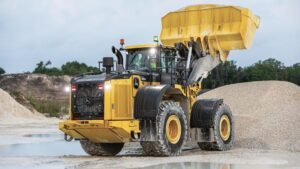 John-Deere-904X-Tier-scaledjpg - Machines Wiki Yellow heavy-duty dump truck dumping a load of gravel at a construction/quarry site with a large gravel pile nearby and a dirt embankment in the background