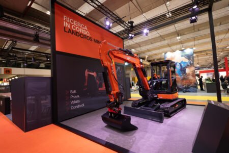 Exhibition booth with a large orange excavator on display inside an industry trade show hall.