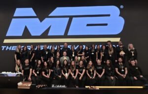 Group of people on a stage posing for a group photo in front of a large blue MB logo backdrop, many wearing black clothing and lanyards.