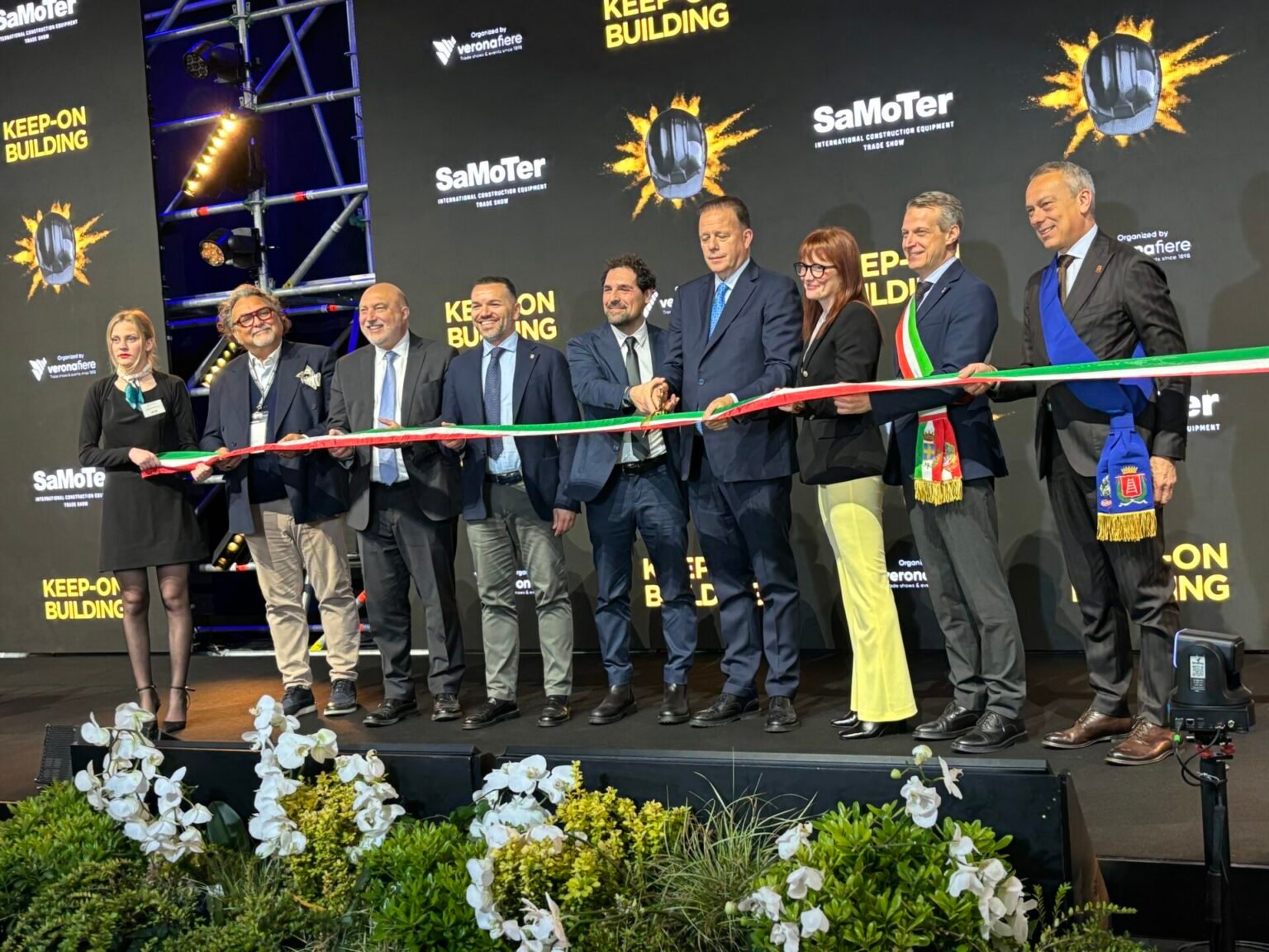 Group of professionals cutting a red-green-white ribbon at a stage ceremony for SaMoTer Keep-On Building, with a black backdrop and floral foregrounds.