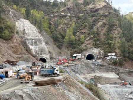 Construction site at a hillside with two tunnel openings under active work; trucks, cranes, and steel bars scattered around the area.