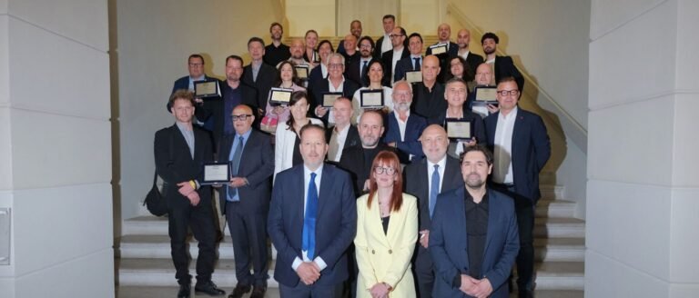 Large group of professionally dressed people posing on a stairway, each holding award plaques at an awards ceremony.