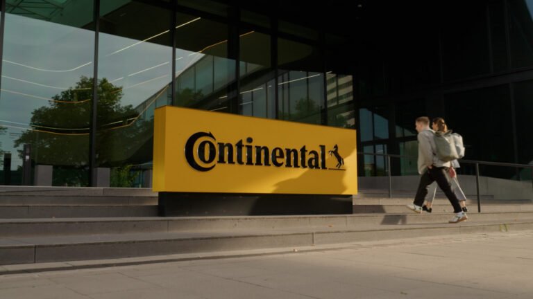 continental_unternehmenszentrale__2025-16_jpg - Machines Wiki Yellow Continental sign with black logo in front of a glass-front building; two people with backpacks walking up the steps nearby.