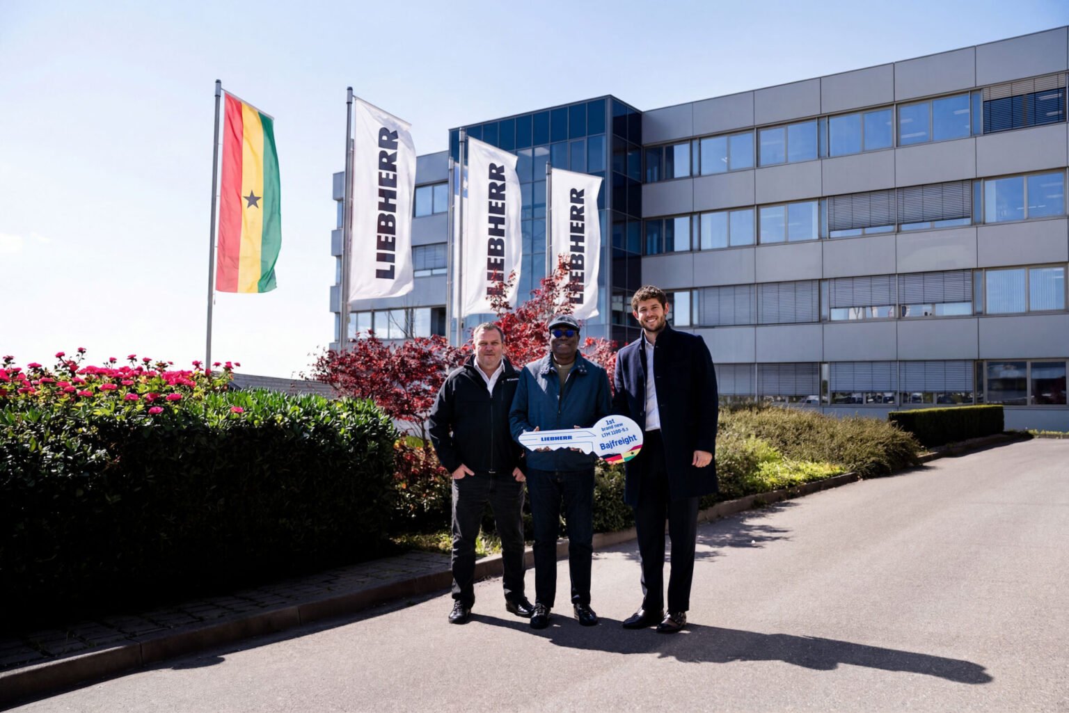 Three men standing outside a modern office building with flagpoles; one man holds a large ceremonial Liebherr key and smiles at the camera.