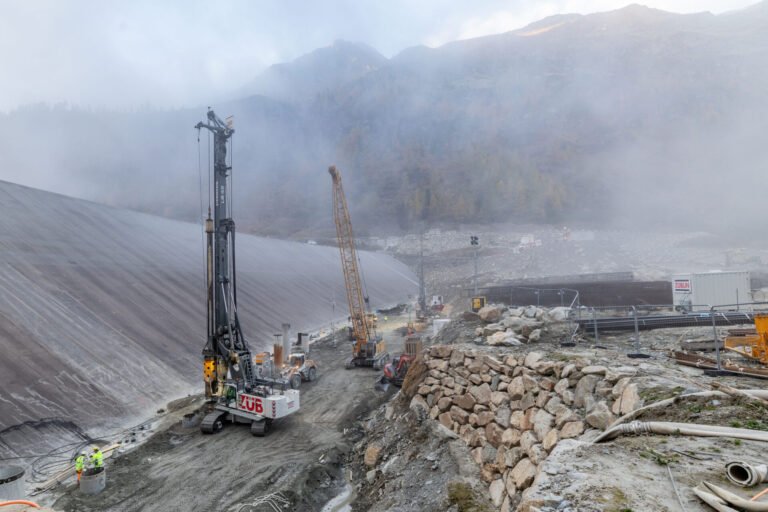 Construction site with a large drilling rig and crane on a misty mountainside; workers in high-visibility gear nearby amid rocky terrain.